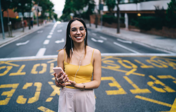 ragazza hipster felice in cuffia che si gode la musica preferita in piedi sulla strada urbana senza traffico, ritratto a mezza lunghezza di gioiosa ragazza millennial che ascolta playlist audio dal moderno dispositivo smartphone - spanish culture audio foto e immagini stock