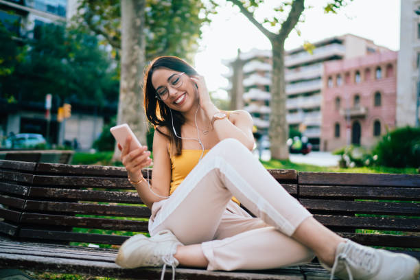 gioiosa ragazza hipster millenaria in auricolari che sceglie la musica audio dall'applicazione per smartphone con playlist, blogger femminile divertente che ascolta canzoni popolari che si godono il tempo in città per la messaggistica di chat - spanish culture audio foto e immagini stock