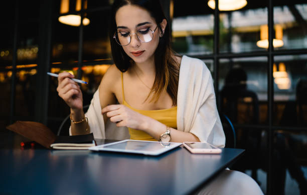 caucasian blogger in glasses installing gadget application for online messaging and chatting, millennial hipster girl 20 years old booking tickets via touch pad during web networking in street cafe - women spain 20 25 years adult imagens e fotografias de stock