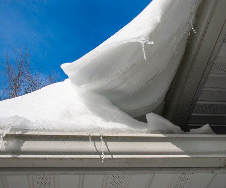Multiple frozen winter icicle stalactites hanging over the edge of the overflowing roof gutters along the eaves and drainage downspouts of a suburban Rochester, New York State residential district house.