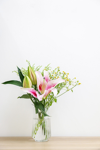 Beautiful bouquet of flowers, a blossoming pink lily, two unopened bulb-shaped lilies, and small daises, in a glass vase, decorating left side of wooden desk against white wall