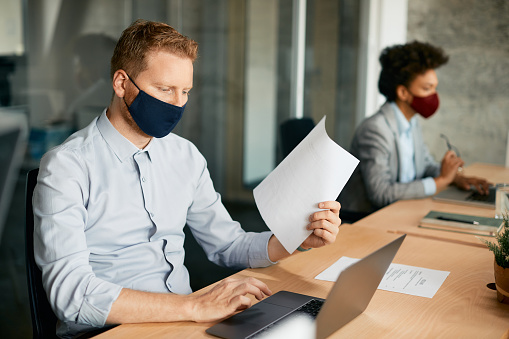 Businessman using laptop while working at corporate office and wearing protective face mask due to coronavirus pandemic.