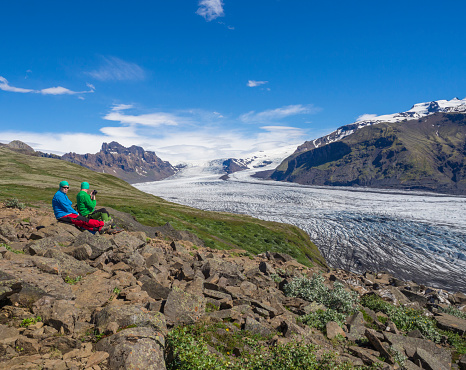 Iceland, Skaftafell national park, July 5, 2018: tourist couple looking at view on glacier lagoon with icebergs and tongue of Skaftafellsjokull, Vatnajokull spur and colorful snow covered rhyolit mountains