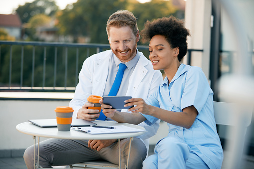 African American nurse and male doctor using mobile phone while having coffee break on a terrace at the hospital.