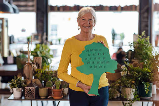A portrait shot of a senior caucasian woman behind the counter of a small plant store. She is looking and smiling at the camera as she holds a cutout of a tree with a tally of how many trees her business has planted.
