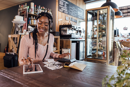 A medium shot of a black woman wearing casual clothing in a cafe in Newcastle Upon Tyne. She is sitting at a table, using a digital tablet and adding up receipts on a calculator.