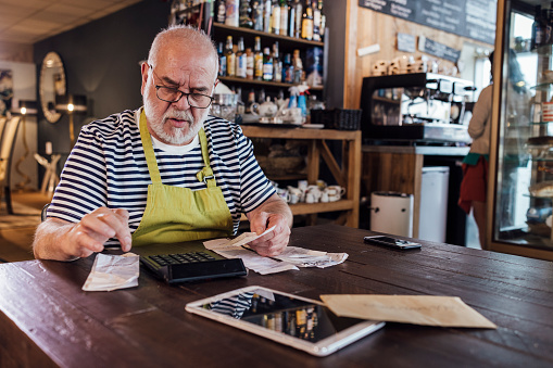 A medium shot of a mature caucasian man wearing casual clothing, an apron and eyeglasses. He is sitting at a table in his cafe adding up receipts on a calculator.