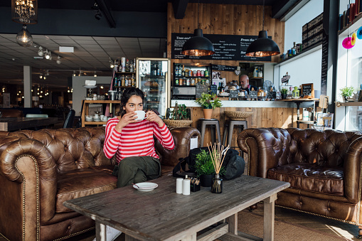 A wide shot of a multiracial woman wearing casual clothing in a cafe in Newcastle Upon Tyne, Northeastern England. She is sitting on a sofa and enjoying a coffee.