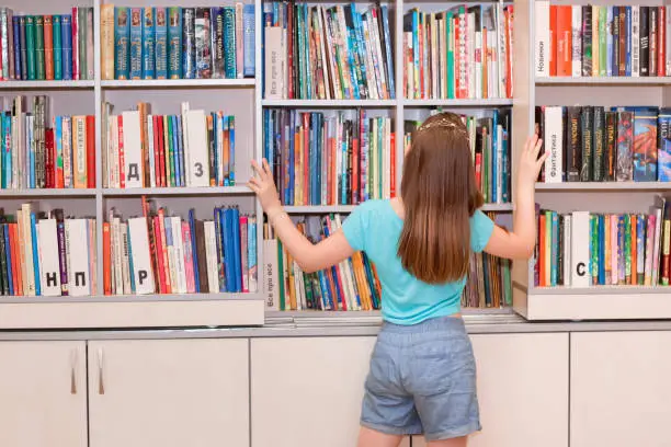 Girl in the library. The girl chooses a book to read. The girl is standing near the bookshelf.