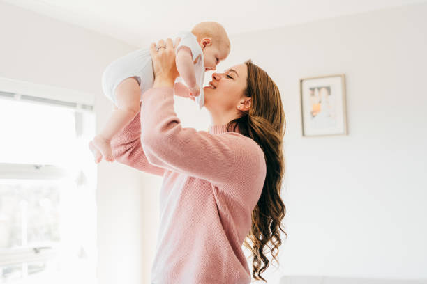 giovane madre con le braccia alzate che tiene il bambino vicino al viso nel soggiorno - love enjoyment innocence clothing foto e immagini stock