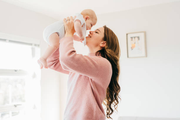 Young mother with raised arms holding baby close to face in living room Proud young mother holding up baby close to face and smiling in living room mother stock pictures, royalty-free photos & images