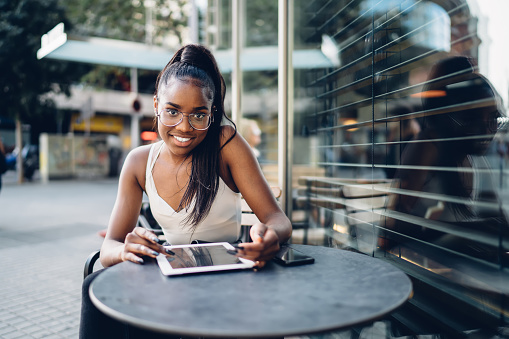 Half length portrait of cheerful african american in eyewear browsing on digital tablet sitting on cafe terrace, young attractive 20s dark skinned woman looking at camera using touchpad on free time