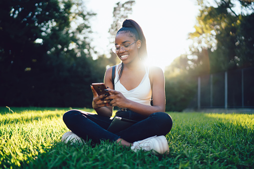 Positive young black lady in casual clothes sitting on green lawn in lotus pose in park and browsing smartphone in daylight