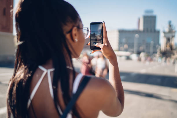 Rear view of dark skinned female holding mobile phone shooting video for share in vlog standing on city square, back view of african american woman blogger taking photo during trip explore locations vista trasera de la mujer de piel oscura sosteniendo el teléfono móvil grabar video para compartir en vlog de pie en la plaza de la ciudad, vista posterior de la mujer afroamericana blogger tomando fotos durante el viaje explorar lugares