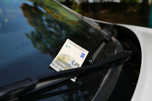 A European Euro banknote on the front windshield of a white car right under the windshield wiper. On the picture there is a five Euro banknotes.
