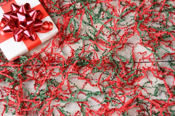High angle view of a single wrapped Christmas present in the upper left corner surrounded by shredded red and green crepe paper.