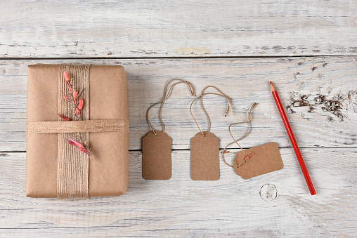 Top view of a Christmas present wrapped with brown paper, burlap ribbon and flowers next to gift tags and a red pencil.