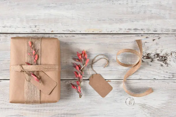 Top view of a Christmas present wrapped with brown paper, burlap ribbon and flowers, next to gift tag flowers and ribbon.