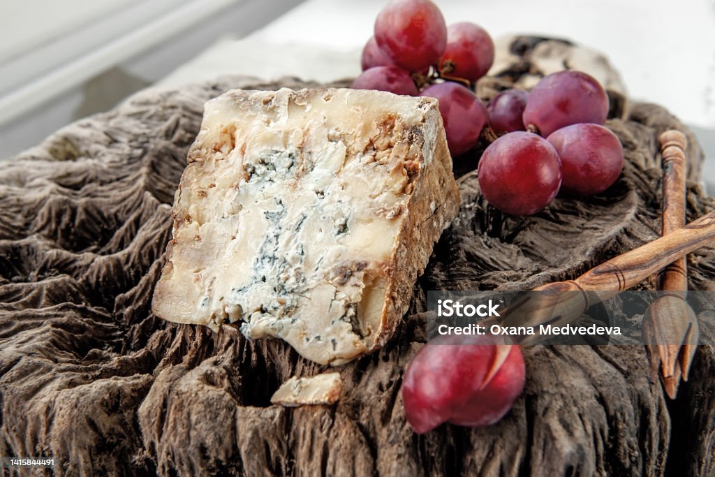 A piece of blue Stilton cheese on a wooden antique background with large red grapes. Close up A piece of blue Stilton cheese on a wooden antique background with large red grapes. Close up. Stilton Cheese Stock Photo