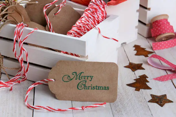 Closeup of a Christmas gift tag leaning on a wood box of wrapping supplies. Horizontal format on a rustic wood table.