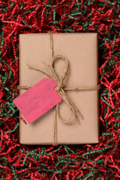 Overhead view of a single Christmas present wrapped with eco friendly craft paper and tied with twine. The package is resting on a field of red and green shredded crepe paper.