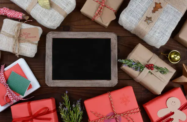 A group of Christmas presents on a dark wood table surrounding a blank chalkboard.