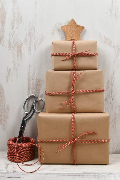 Three plain brown paper wrapped gifts stacked in the shape of a Christmas tree with star. Scissors and ball of twine are next to the packages.