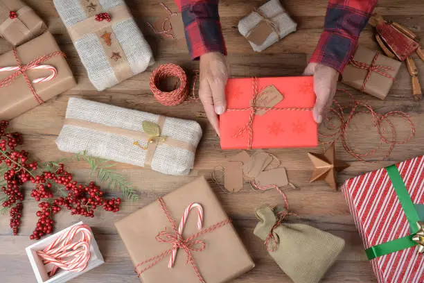 High angle shot of a man holding a Christmas Present over a table filled with wrapped gifts and wrapping supplies.