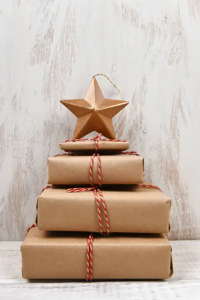 Plain brown paper wrapped gifts tied with twine and stacked in the shape of a Christmas tree with star, against rustic white wood background.