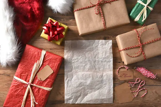 Overhead view of wrapped Christmas presents on a rustic wood table with string, gift tags, and Santa hat. Horizontal format with a blank sheet of paper.