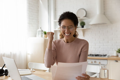 Euphoric Hispanic woman sit in kitchen read paper notification from bank about last loan payment feels happy, get job promotion letter, learn sales growth look excited. Great unbelievable news concept