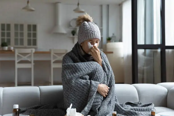 Photo of Sick woman covered with plaid, blowing runny nose in napkin