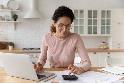 Hispanic woman use calculator calculate costs working sit at table in kitchen. Pay monthly bills through e-bank app on laptop. Business, summarize household expenses, manage budget, accounting concept