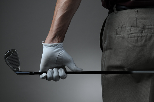 Closeup of a male golfer holding a six iron behind his body. Man has a Golf Glove on his hand. Horizontal format over a light to dark gray background.
