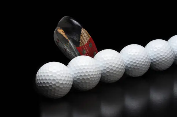 Golf Still Life. An antique golf club behind a line of new golf balls, on black with reflection.