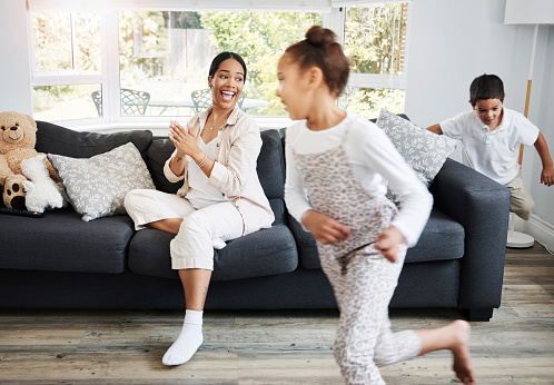 Playing, running and young children excited indoors with a smiling mother watching on a couch. Happy family spending the day together indoors while the active kids run, play and have fun at home