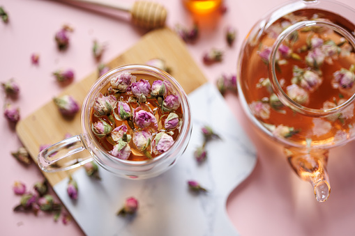 Istanbul, Turkey-August 19, 2022: Herbal tea in a glass cup decorated with dried rose buds on a marble-wood presentation board on a light pink background. Next to the herbal tea is a pot of herbal tea. Rose buds are scattered all around. Double wall glass. Shot with Canon EOS R5.