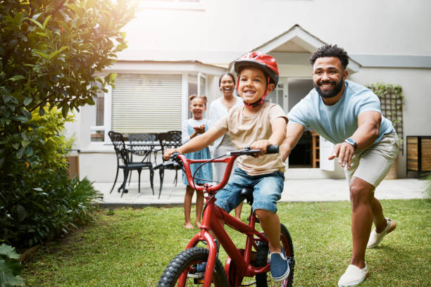 aprendizaje, bicicleta y padre orgulloso enseñando a su hijo pequeño a montar mientras usa un casco por seguridad en el jardín de su casa familiar. padre activo que ayuda y apoya a su hijo mientras anda en bicicleta afuera - happiness smiling offspring family fotografías e imágenes de stock