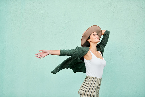 Portrait of a beautiful young woman with her eyes closed holding her hat and arm outstretched while standing in front of turquoise color wall