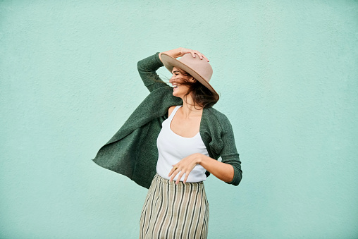 Portrait of a beautiful young woman holding her hat in breeze and looking away smiling while standing in front of turquoise color wall
