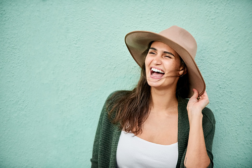 Portrait of beautiful young woman with hat standing against turquoise color wall and laughing