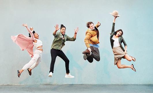 Full length of group of excited men and women friends in stylish wear jumping in air against a light blue wall outside