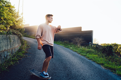 Full length anonymous man skater in back lit standing with on leg on skateboard and looking at wristwatch in countryside in sunset time