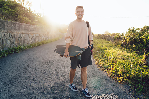 Half length portrait of cheerful caucasian male skater walking on rural asphalt road enjoying summer weekends, handsome hipster guy 20s with board spending free time on active sport hobby outdoors
