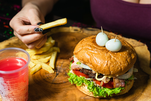 close-up of a latin girl with her hamburger with fries and red fruit juice, served on the wooden table, ready to start eating. woman with a potato in her hand about to put it in her mouth. street food