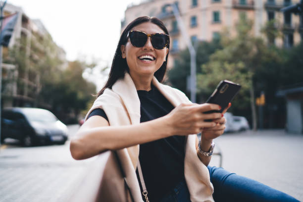 Beautiful trendy dressed female blogger laughing on funny content using mobile phone on free time outdoors, smiling woman 20s enjoying 4G connection on street in roaming chatting via application hermosa bloguera vestida de moda riendo de contenido divertido usando el teléfono móvil en el tiempo libre al aire libre, mujer sonriente de 20 años disfrutando de la conexión 4g en la calle en roaming chateando a través de la aplicación