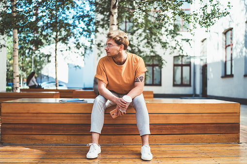 Serious man in glasses and stylish clothes holding disposable cup of fresh coffee and looking away while chilling on wooden bench
