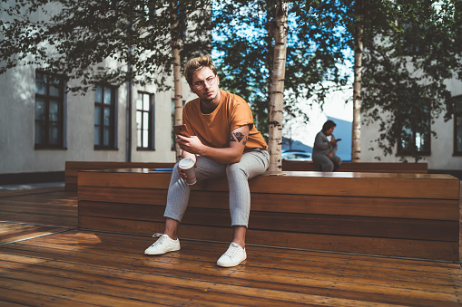 Focused man in glasses and stylish clothes sitting on wooden bench with cup of coffee and notebook holding mobile phone in hand