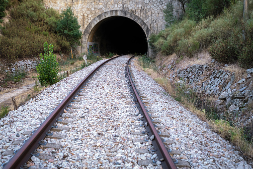 entrance of a tunnel on a railroad track, close up of the rails forming a curve before entering into the darkness of the tunnel, vegetation on the sides, point of view