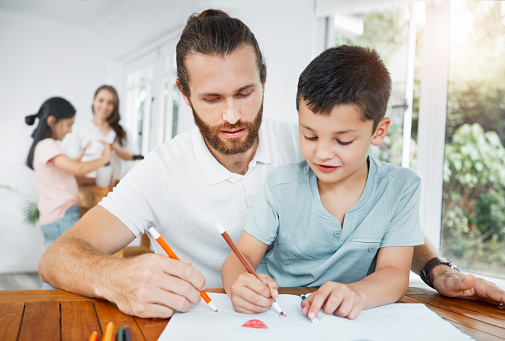 Father and son bonding, drawing and spending time together with the family in the background at home. Little boy and his male parent coloring in and having fun while doing homework and learning